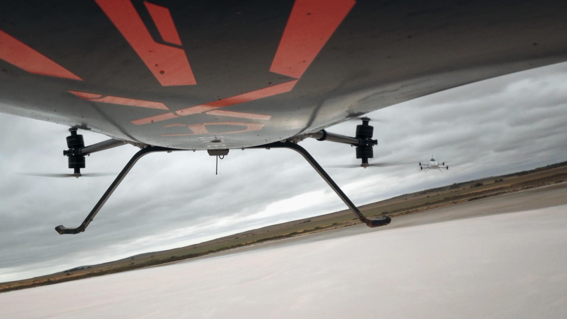 Close-up view of a drone hovercraft flying over a flat body of water with another drone visible in the background under a cloudy sky.