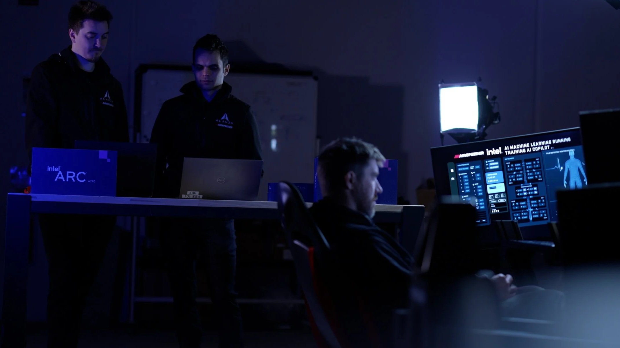 Two Engineers and a Pilot in workshop working at computer stations with large screens displaying AI machine learning data, illuminated by bright lights.