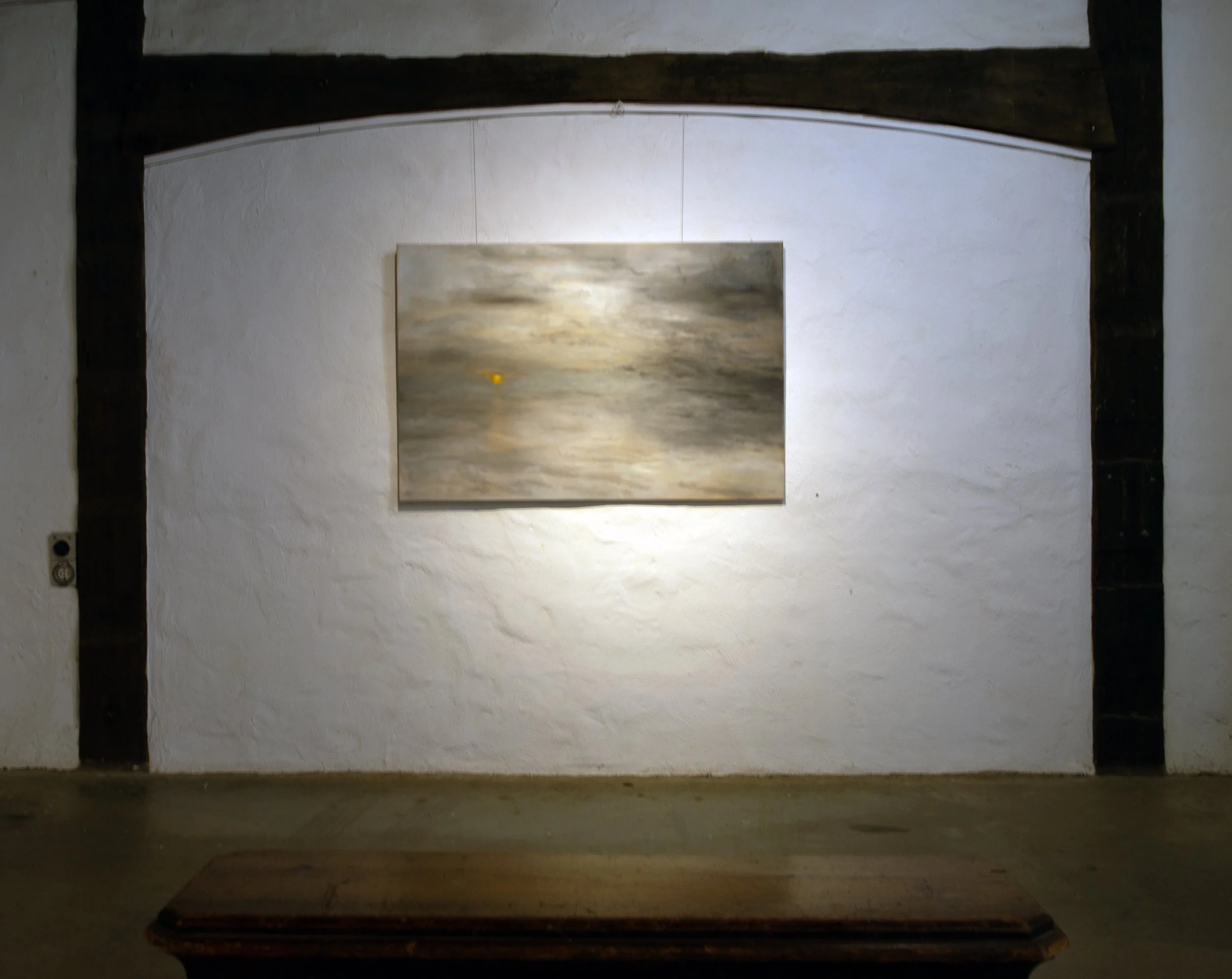 A painting hanging on a white textured wall in a room with wooden beams. The painting depicts a cloudy sky with sunlight breaking through and a small yellow object, possibly a bird or a balloon, in the sky.