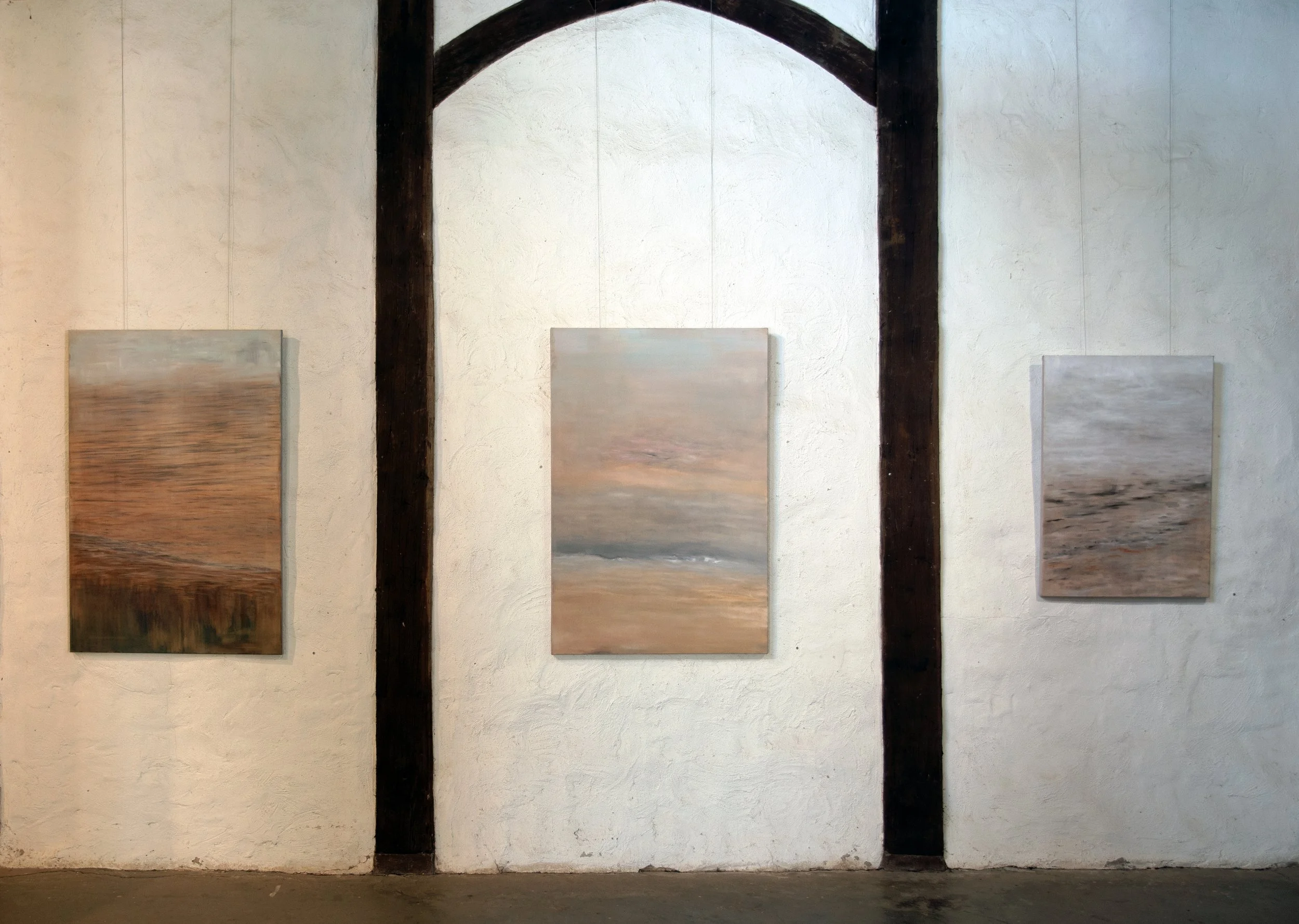 Three paintings that depict a landscape scene with a body of water and sky, displayed against a white wall with dark wooden beams.