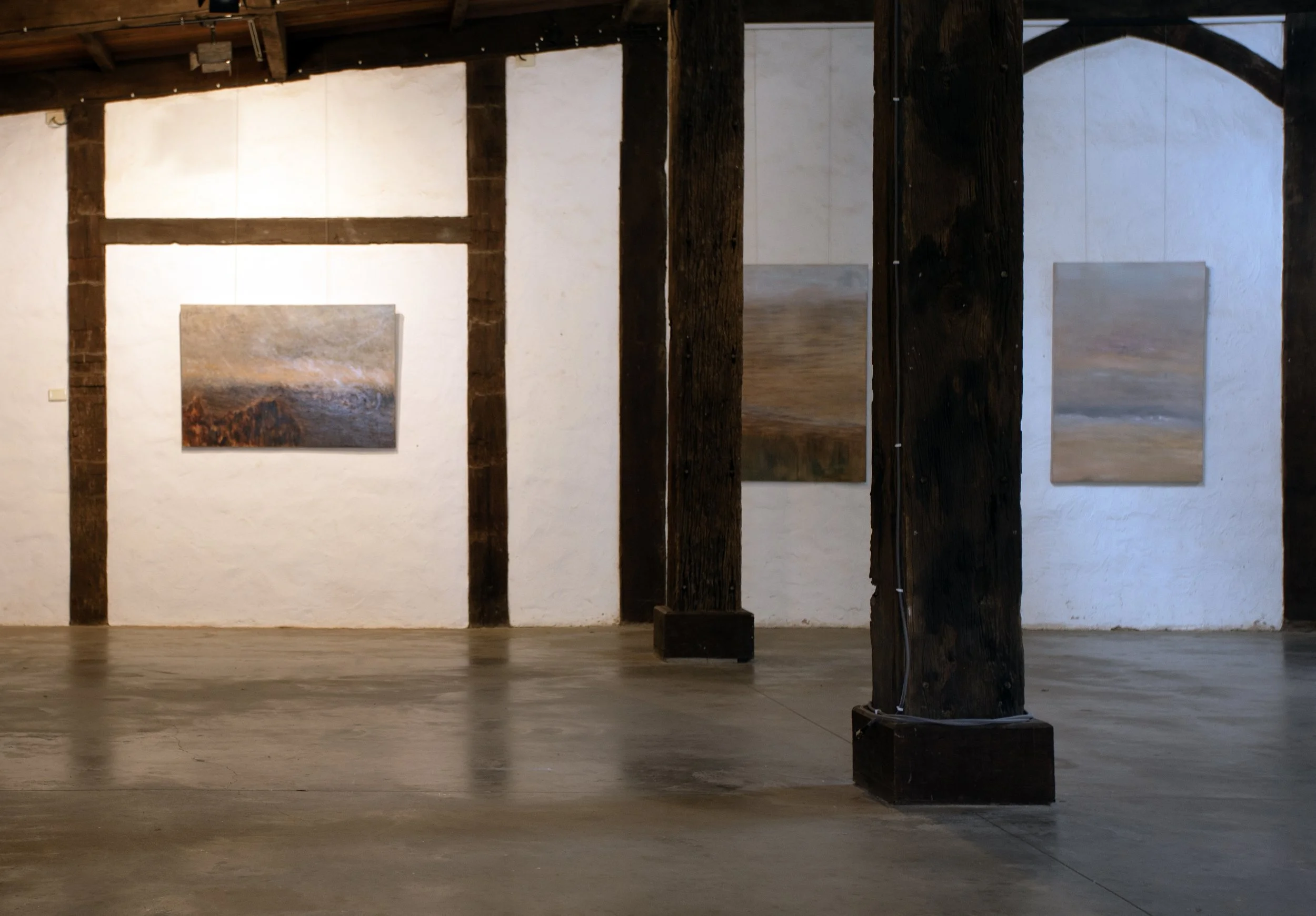 An art gallery with paintings on white walls, wooden beams on the ceiling, and a polished concrete floor. Wooden columns support the structure.
