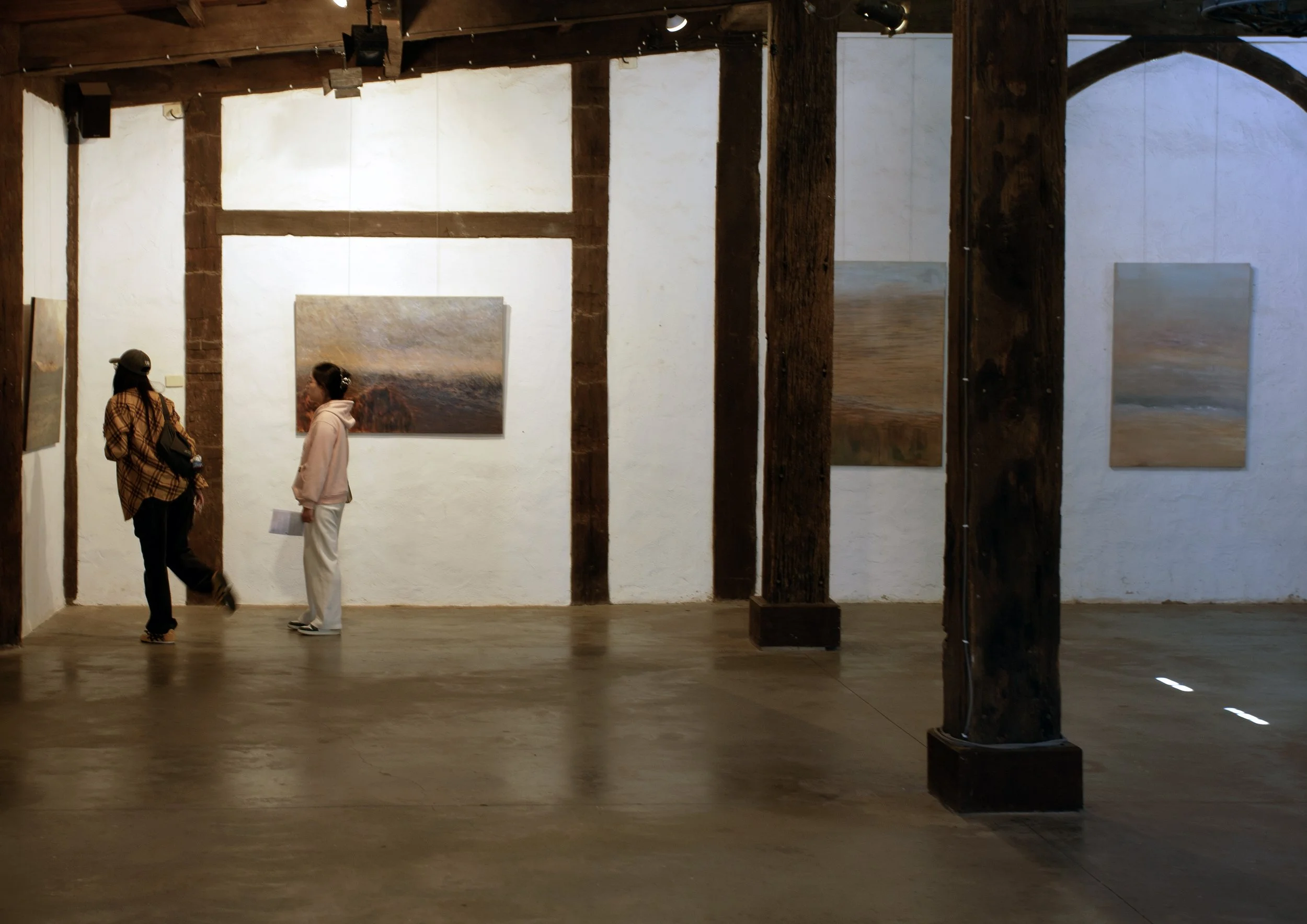Two women standing in an art gallery, looking at paintings on the wall. The gallery has wooden beams and posts, with a polished concrete floor.