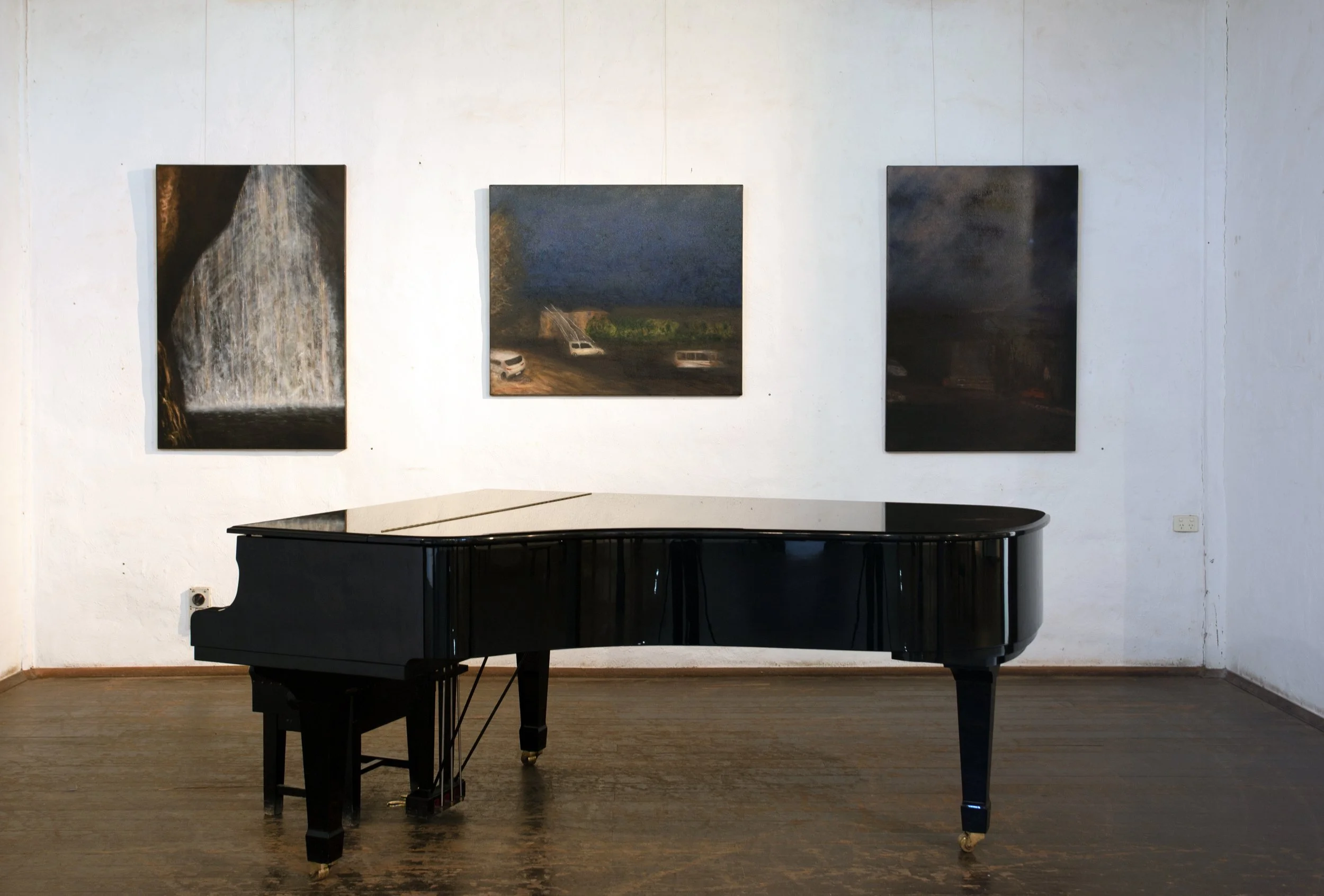 A black grand piano in a gallery or museum with three landscape paintings hanging on a white wall behind it.