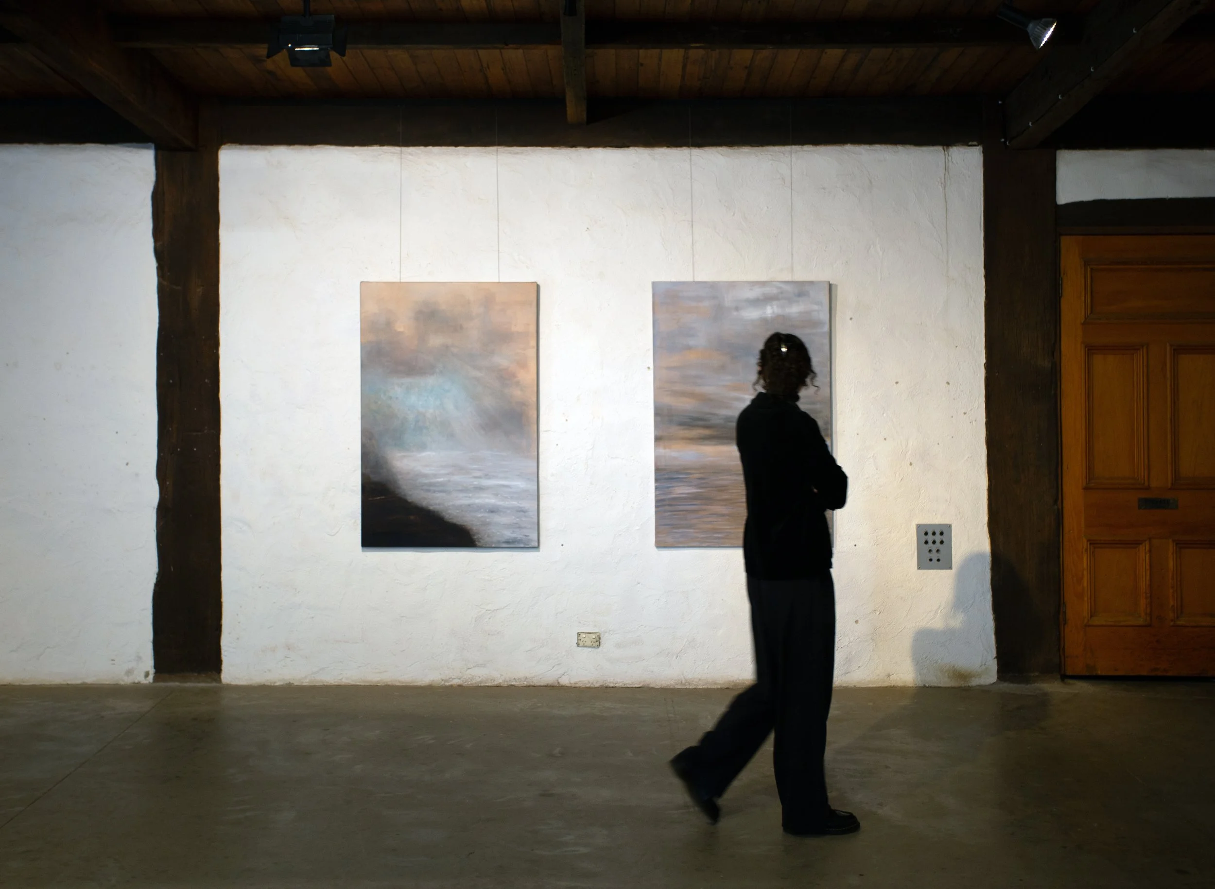 Person with dark curly hair and black clothing walking inside an art gallery with paintings on the white wall.
