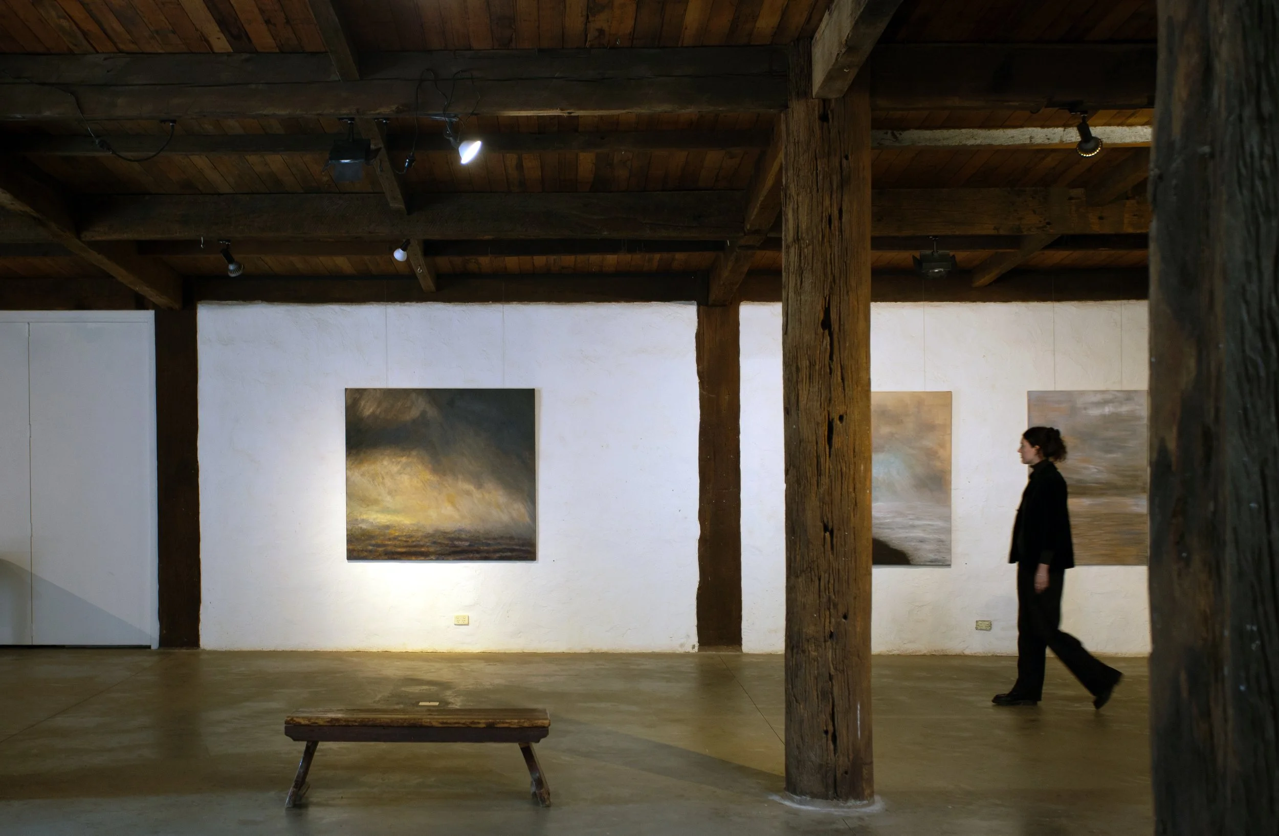 An indoor art gallery space with wooden ceiling beams and white walls featuring paintings, with a person walking past in black attire.