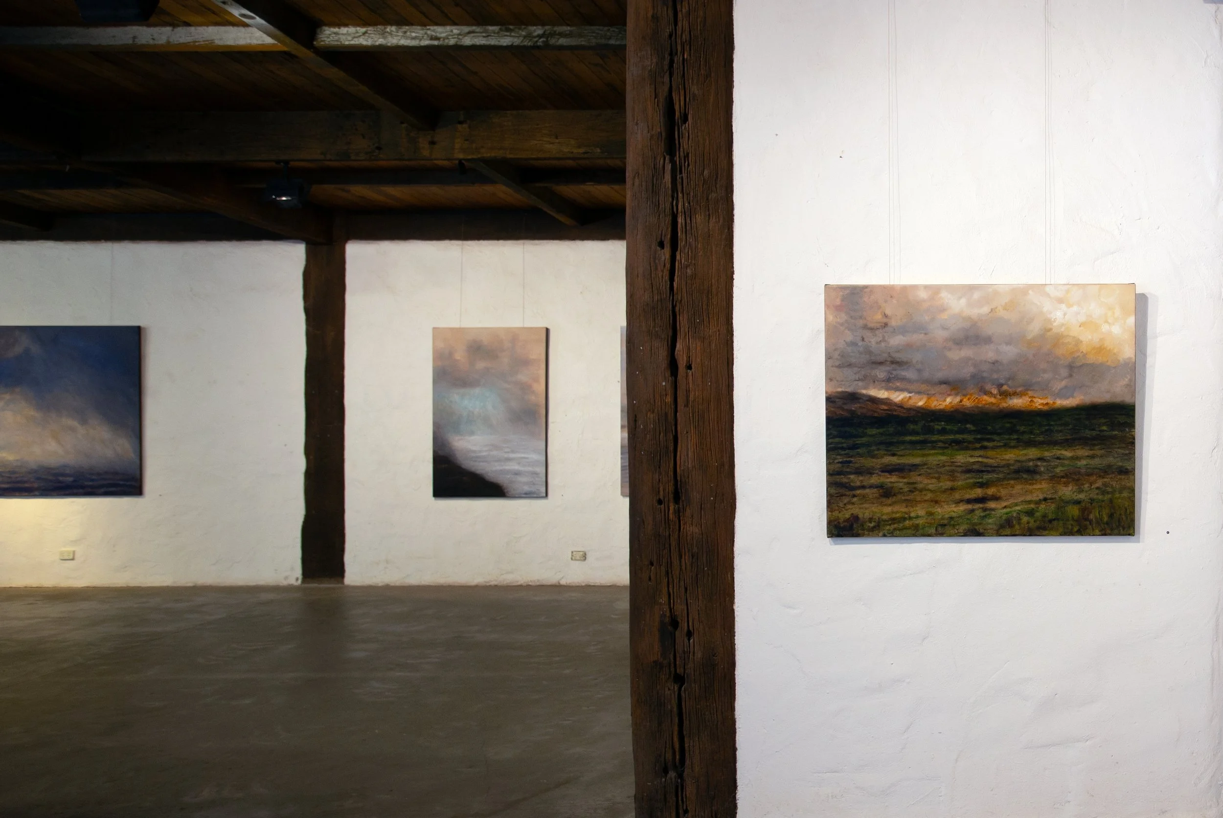 Photo of an art gallery with landscape paintings on white walls and a wooden beam in the center of the room.