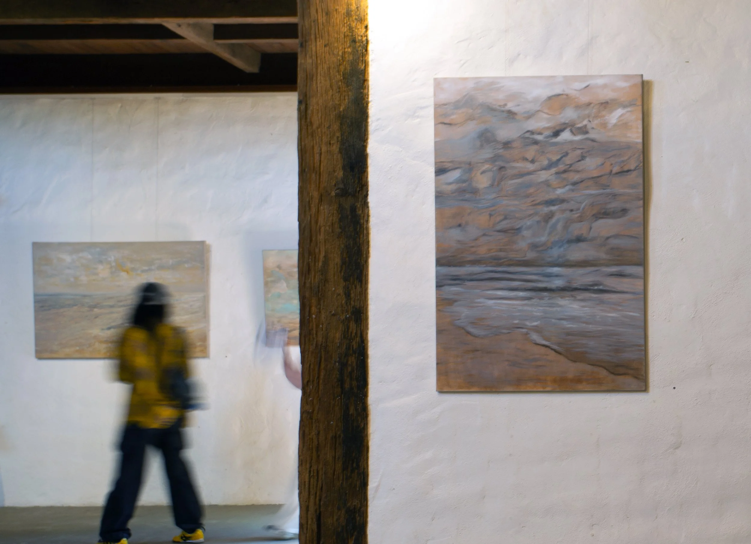 An art gallery with abstract landscape paintings on white walls and a wooden beam in the center. A blurry figure of a person wearing a yellow jacket and black hat is walking past.
