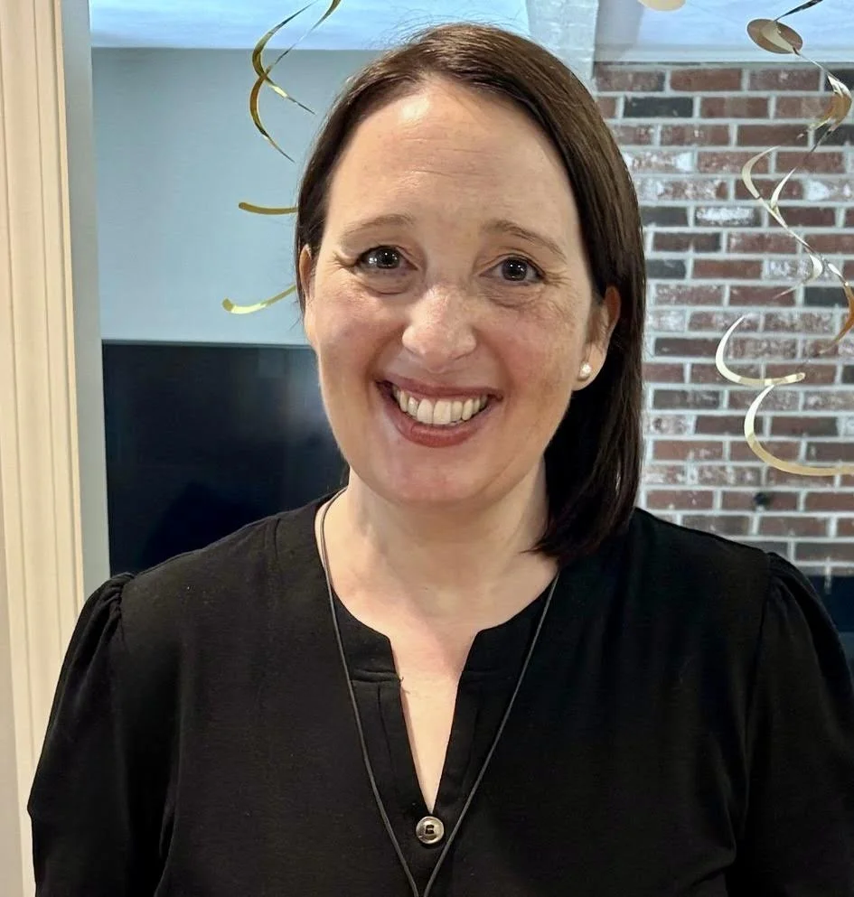 A woman smiling indoors, wearing a black blouse with a necklace, standing in front of a brick wall and gold hanging decorations.