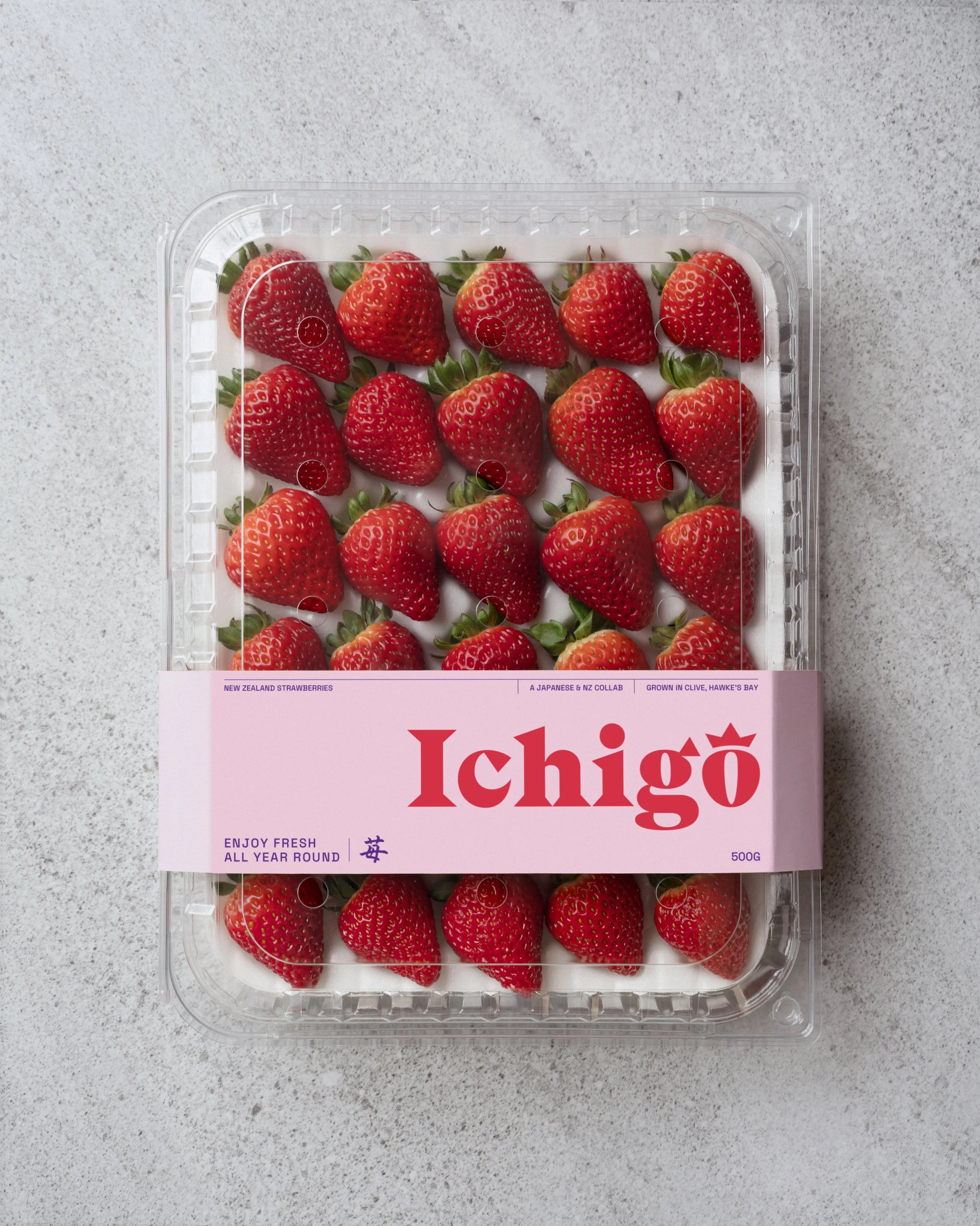 A  container filled with fresh strawberries on a gray speckled surface, with a pink label that reads "Ichigo" and mentions New Zealand strawberries and other details.