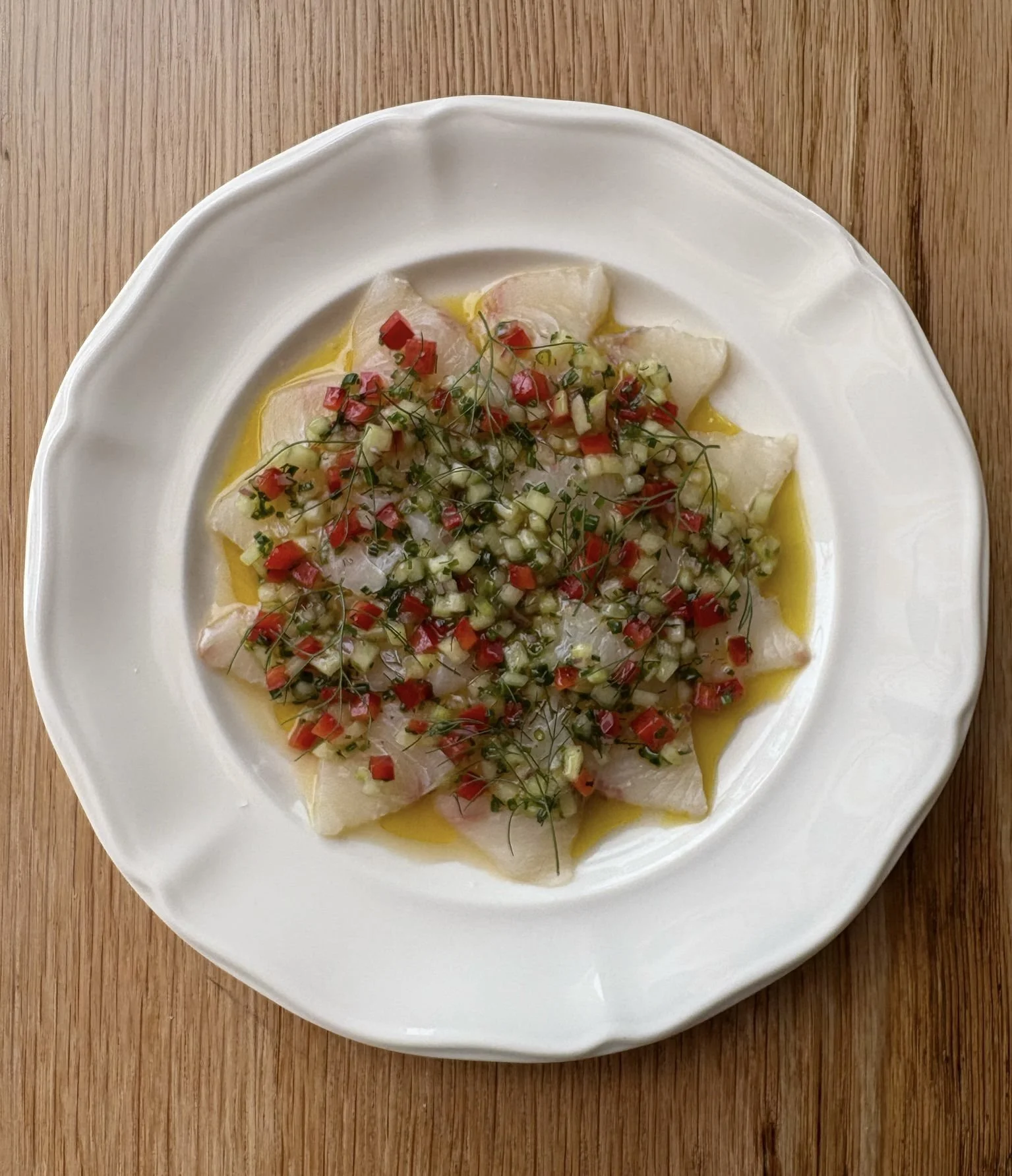 Thin slices of white fish topped with chopped red peppers, green herbs, and diced vegetables, served on a white plate with olive oil, on a wooden surface.