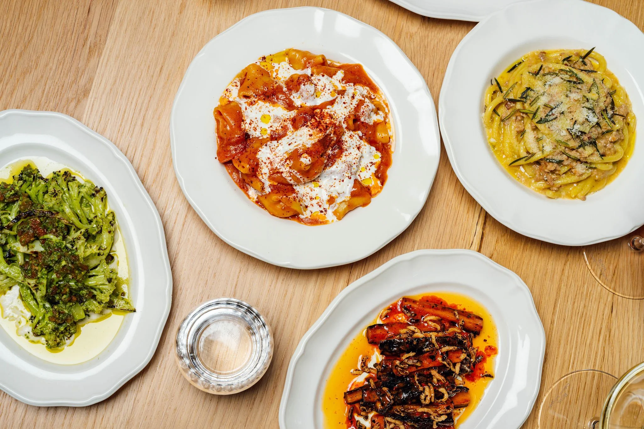 Top-down view of four plates of pasta dishes on a wooden table, including green pesto pasta, flat ravioli with tomato sauce and cream, linguine with meat sauce and parmesan, and pasta with tomato sauce and vegetables.