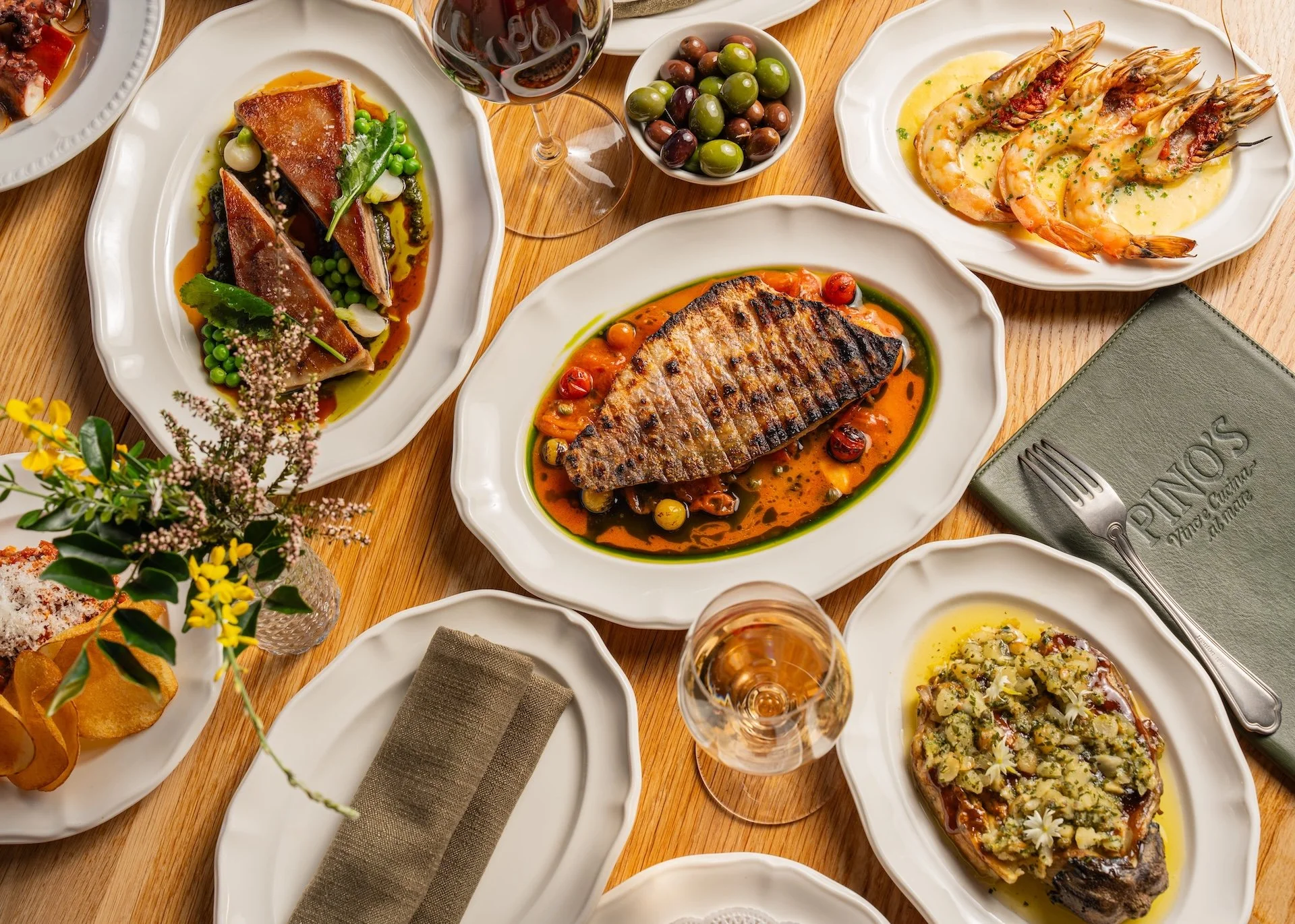 A variety of plated gourmet dishes including grilled fish with cherry tomatoes, shrimp with butter sauce, and other carefully prepared gourmet foods on white plates, with glasses of rosé wine on a wooden table.