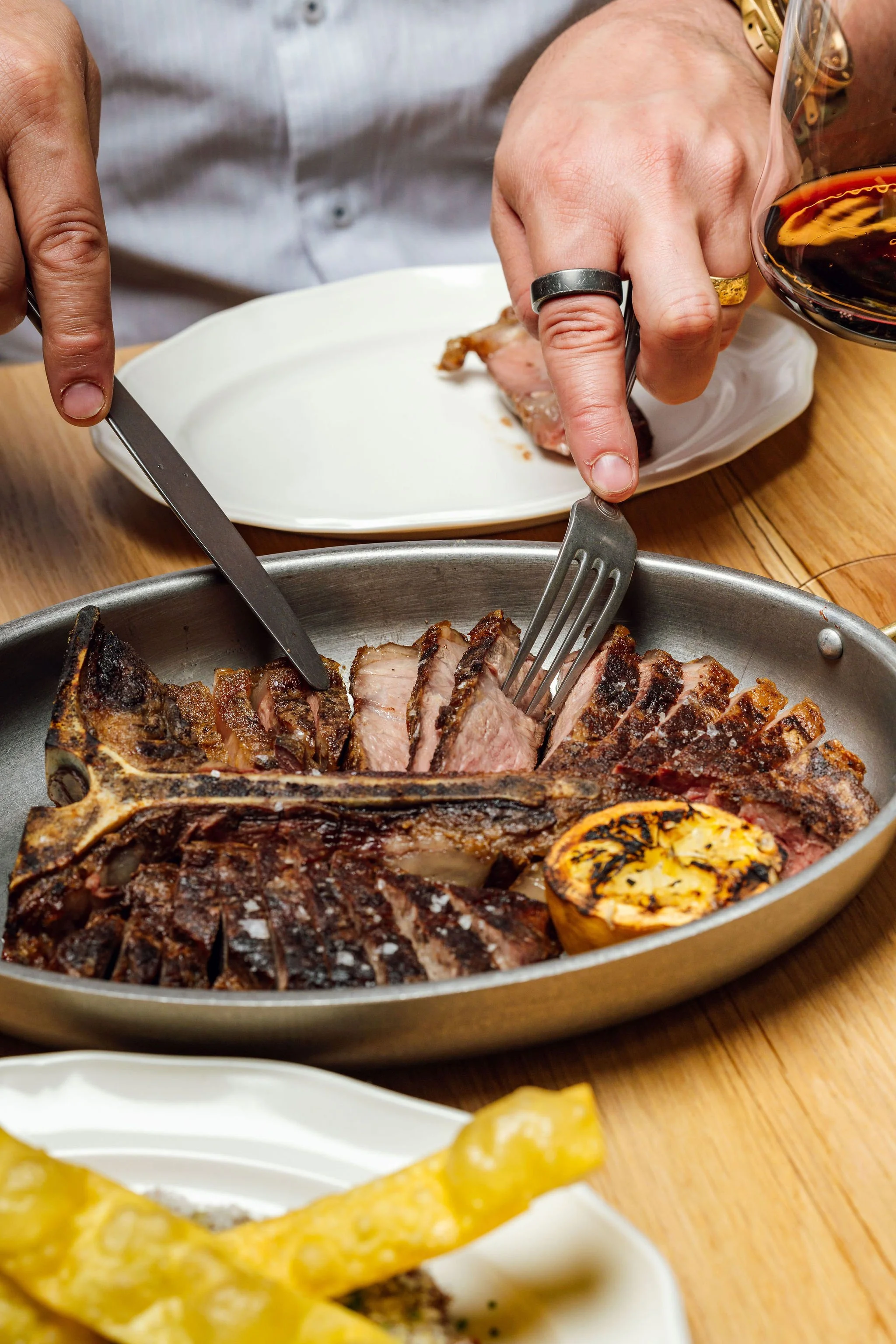 Person cutting into a cooked steak with a fork and knife at a dinner table, with a glass of red wine nearby.