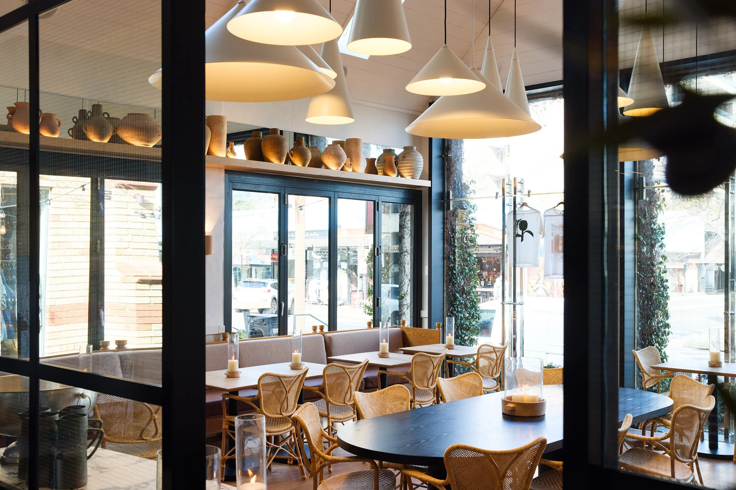 Inside a cozy restaurant with wooden tables, wicker chairs, and candle decor, illuminated by cone-shaped pendant lights, with large windows and decorative pottery displayed on a shelf.