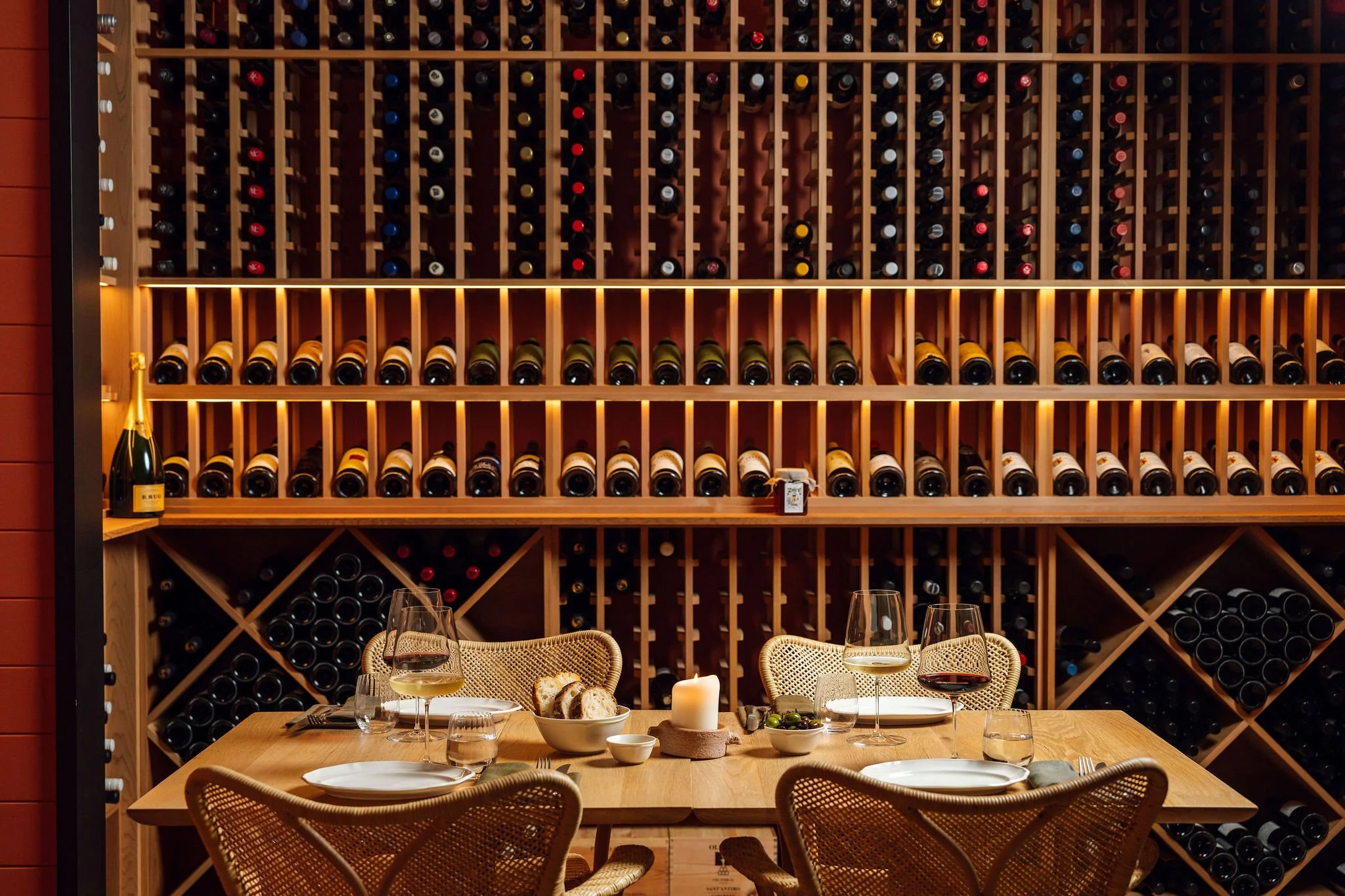 A dining table set for four with wine glasses, bread, and a lit candle in front of a wall of wooden wine racks filled with bottles in a restaurant or wine cellar.