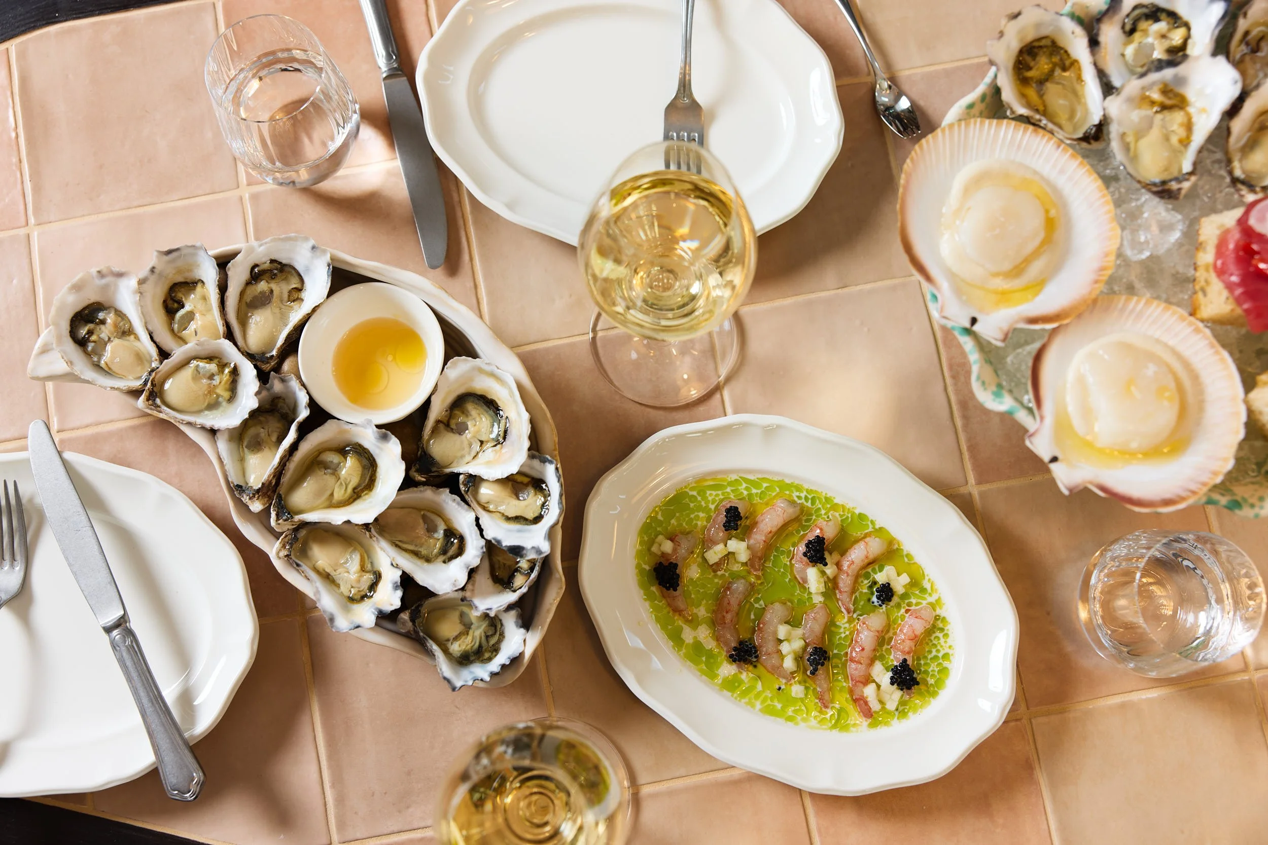 A table with oysters on a platter, a seafood dish with shrimp in green sauce, scallops, and glasses of white wine and water.