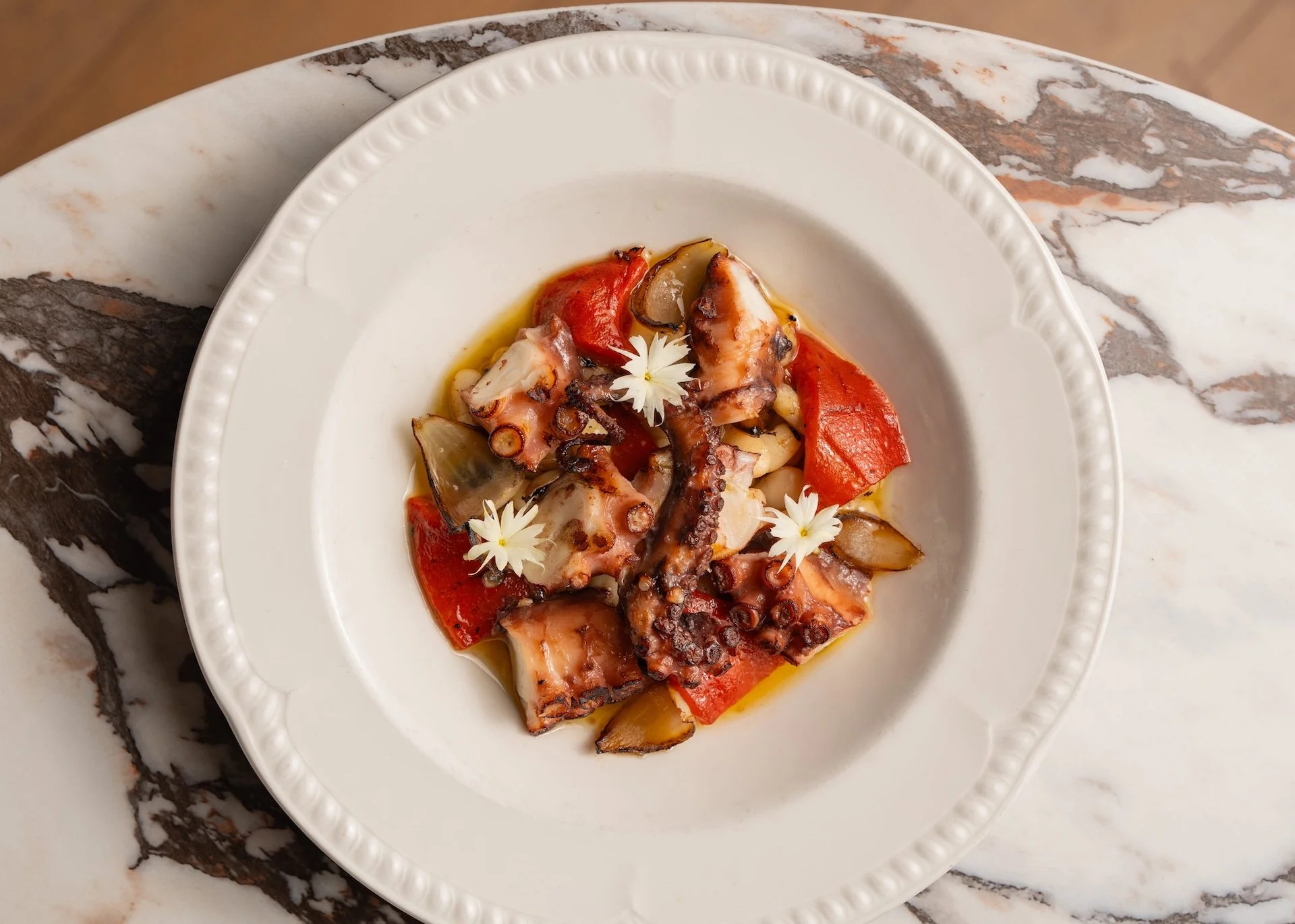 A plate of grilled octopus with red bell peppers, eggplant, and white edible flowers on a marble surface.