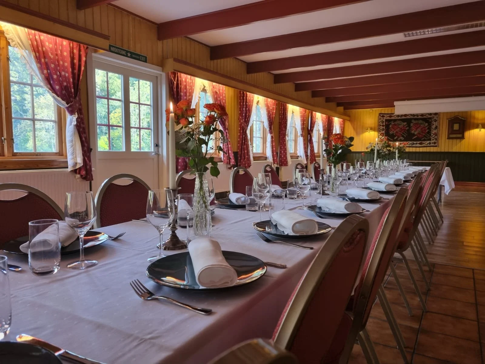 Elegant langbord dekket for et selskap med blomsterhåndtak, lys, hvite duker, vin- og vann glass, tallerkener og bestikk i et rustikt rom med store vinduer med røde gardiner.