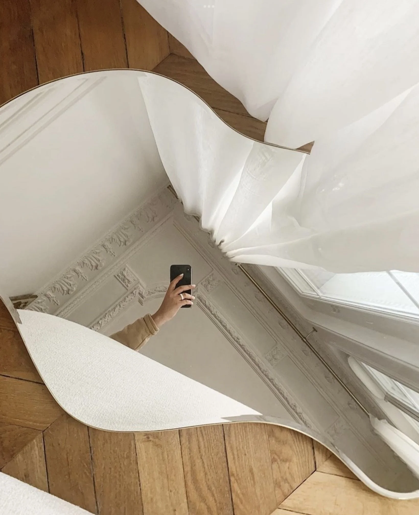 Miroir décoratif en forme de vague placé sur un sol en parquet, reflétant une personne prenant une photo avec un smartphone dans une pièce aux murs ornés de moulures classiques.