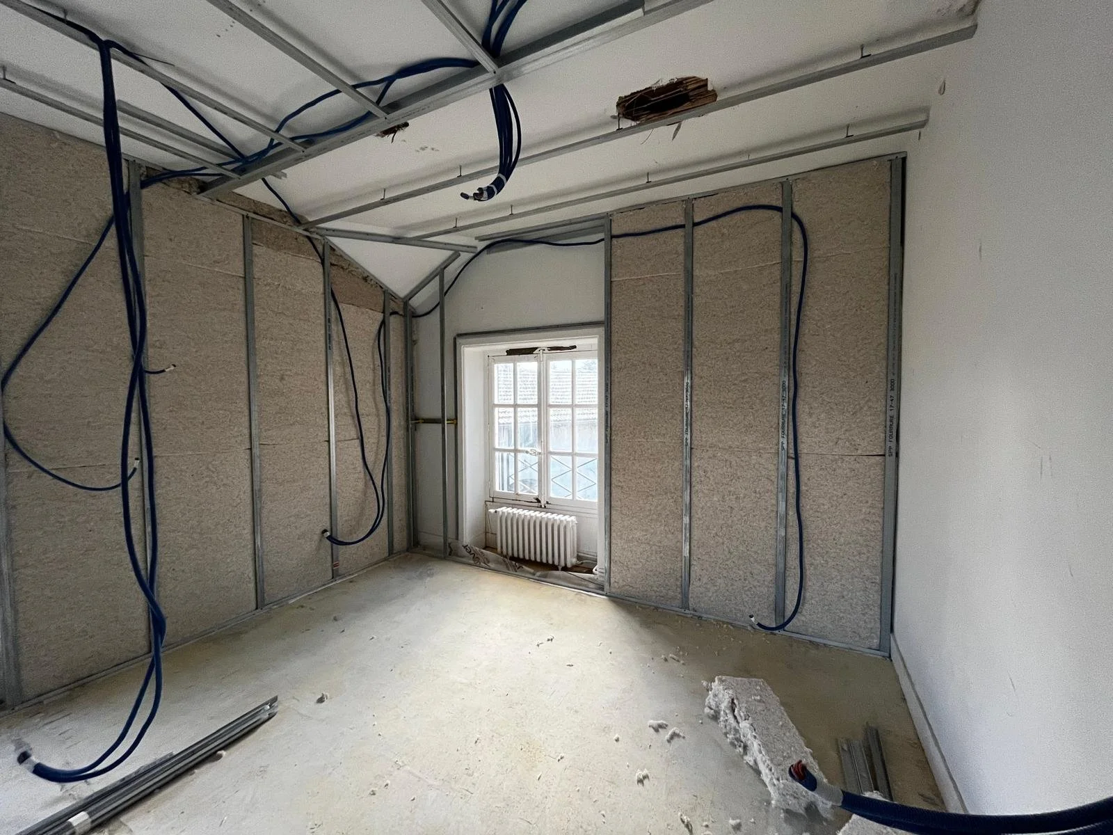 Pièce en rénovation avec ossature métallique, panneaux isolants, câbles électriques apparents, fenêtre et radiateur