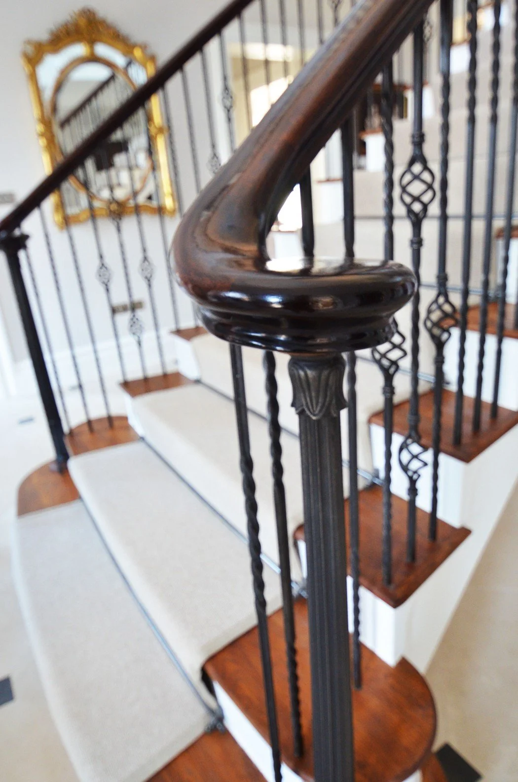Close-up of an elegant staircase with a polished wooden handrail and ornate metal balusters. A decorative mirror with a gold frame is visible on the wall in the background.