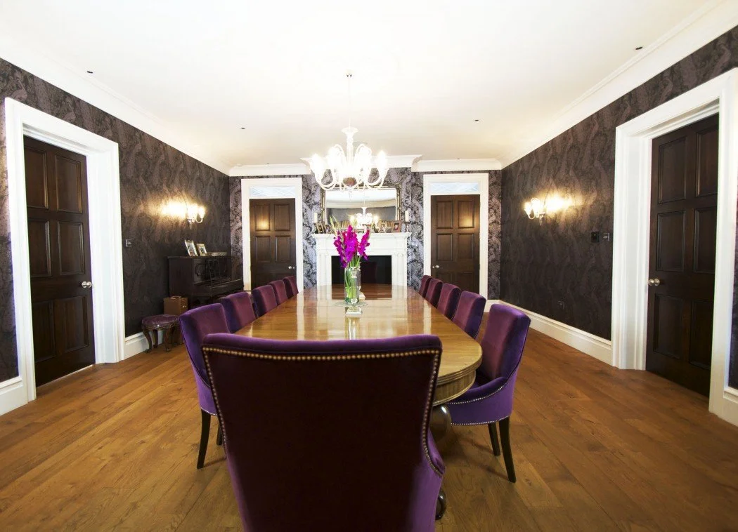 Elegant dining room with a long wooden table surrounded by purple chairs, a chandelier overhead, and a flower vase as the centerpiece. The room features dark wood doors, patterned wallpaper, and a fireplace with decorative items.