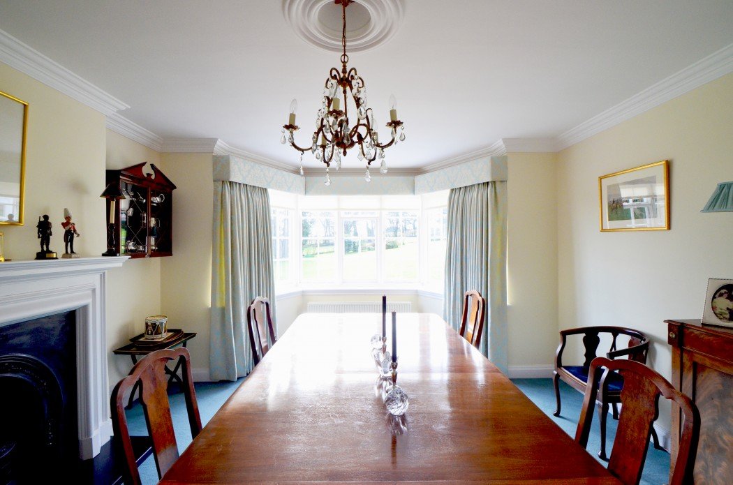 Elegant dining room with wooden table and chairs, chandelier, large window with curtains, light walls, decorative fireplace, and artwork.