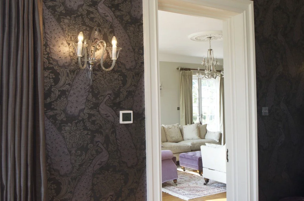 Interior showing a room with dark patterned wallpaper, wall sconce, and view into a living area with a sofa, purple ottoman, and chandelier.