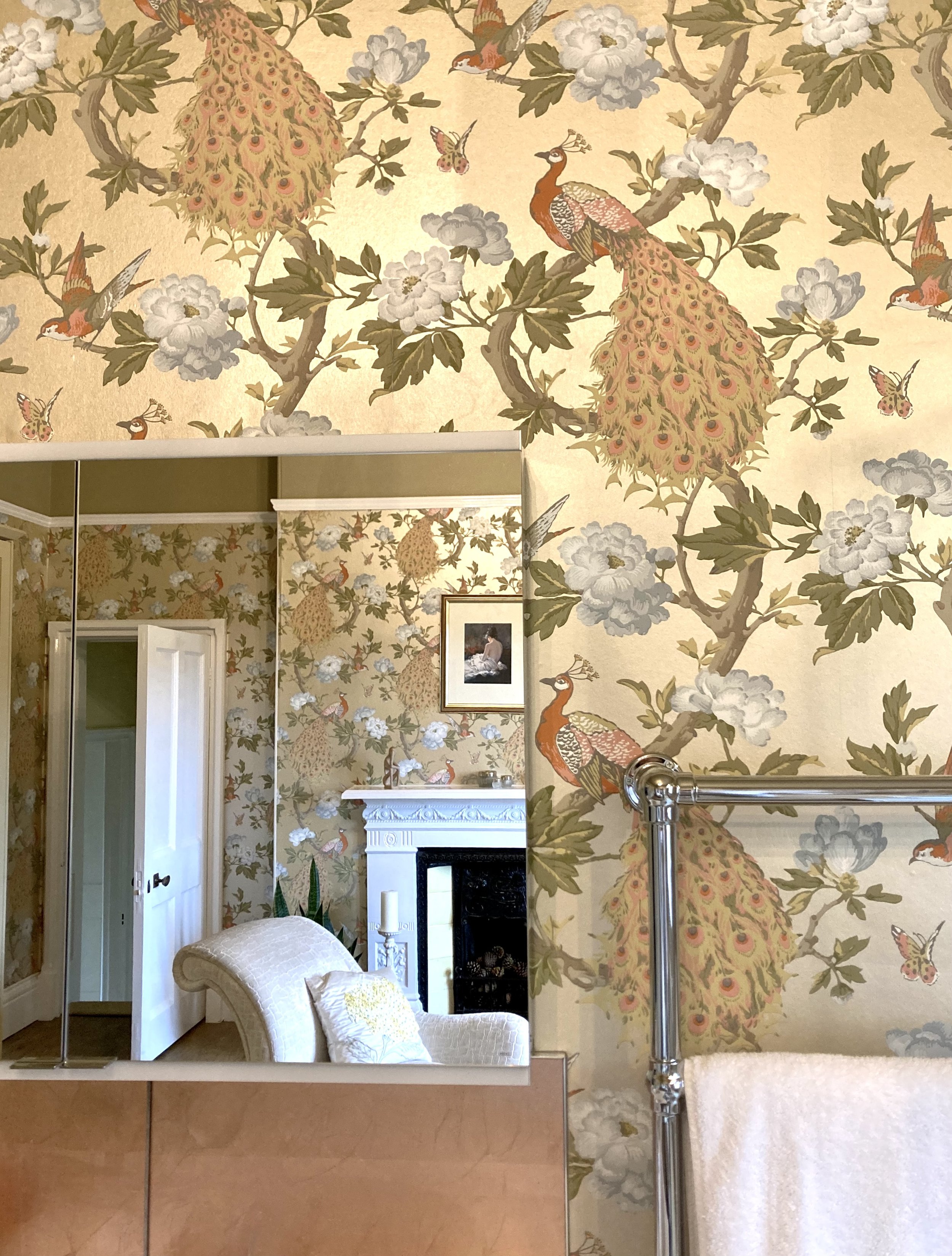Room with floral and peacock wallpaper, wall mirror, and white upholstered chair.