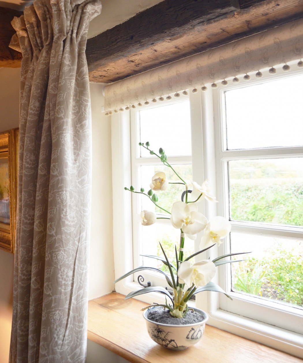 A potted orchid with white flowers on a windowsill, next to patterned curtains and a wooden beam, with a countryside view outside the window.