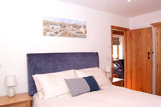 Cozy bedroom with a bed featuring white bedding, a blue headboard, and a patterned pillow. Wooden nightstands with lamps are on either side, and a wooden wardrobe is visible. A landscape painting hangs on the white wall.
