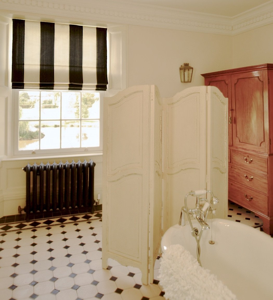 Elegant bathroom with a freestanding bathtub, decorative privacy screen, black and white checkered floor, wooden cabinet, ornate wall molding, black radiator, and striped window shade.