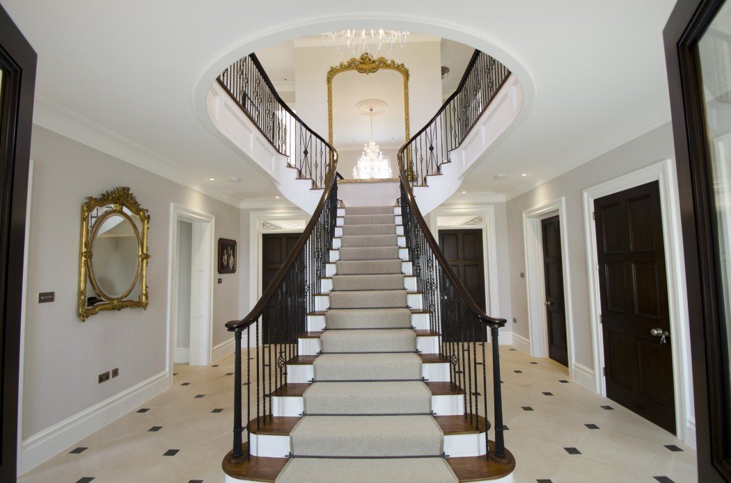 Elegant foyer with a grand staircase, featuring a carpeted center aisle, dark wood handrails, and ornate wrought iron balusters. Chandelier above and a large mirror on the wall.