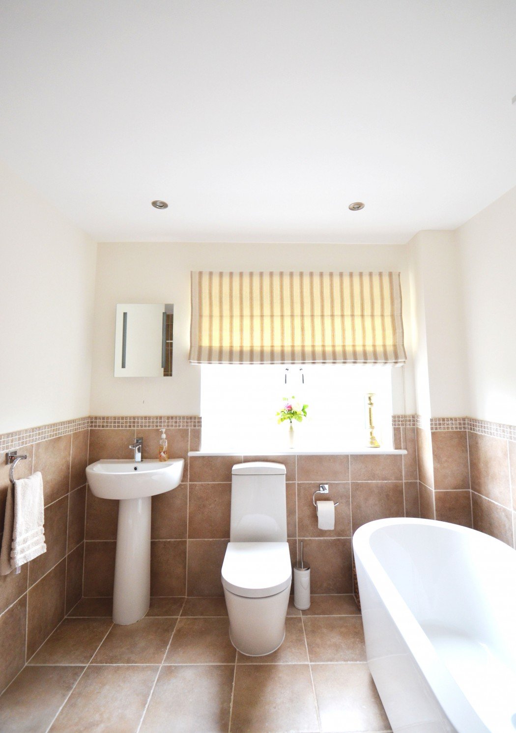 Modern bathroom with a pedestal sink, toilet, bathtub, beige tiles, and a striped window blind.