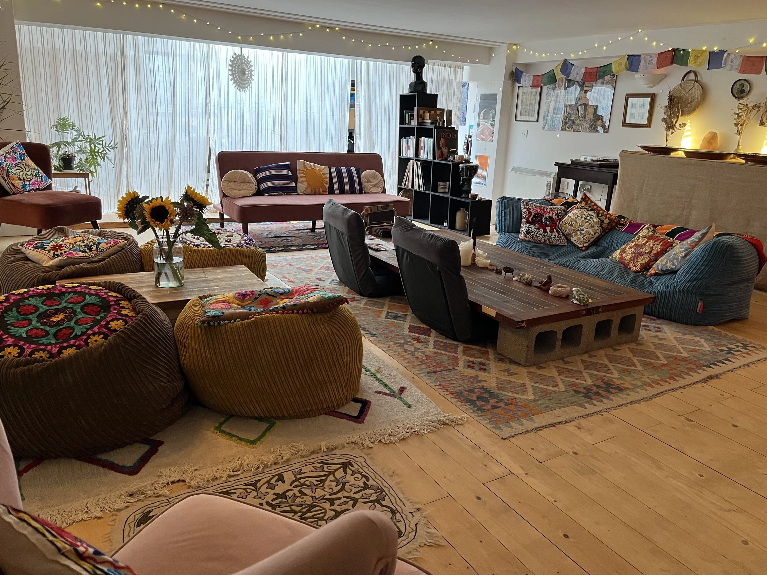 A cozy living room with colorful, eclectic decor. There are multiple sofas and chairs, including a blue sofa with patterned pillows, a pink loveseat, and an orange and brown striped chair. A wooden coffee table is in the center, decorated with shells and stones. A vase of sunflowers is on a side table. Sunlight filters through large sheer curtains, creating a warm atmosphere. The room features various wall art, a shelf with books and decorative items, string lights, and cultural decorations like prayer flags.