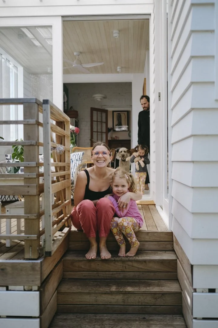 Woman and child sitting on wooden steps, smiling with another child, an adult, and a dog in the background inside a house.