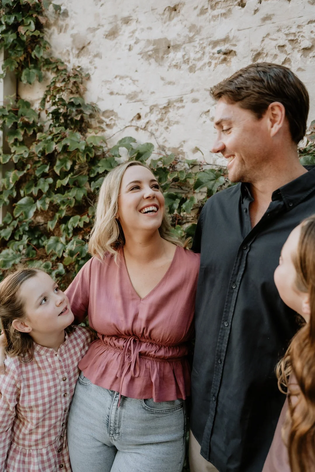 A smiling family of four looking at each other in front of a stone wall with ivy. The woman in a pink blouse stands next to a man in a black shirt, with two children beside them.