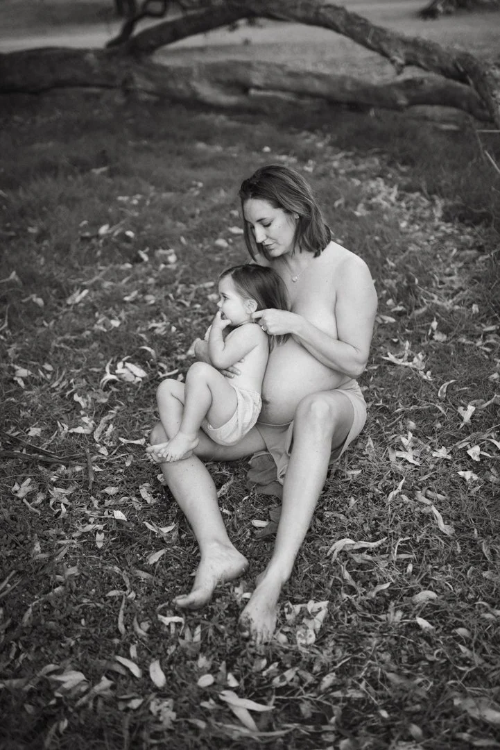 A pregnant woman sitting on grass, holding a young child on her lap, both looking content and relaxed. The background features natural scenery and fallen leaves.