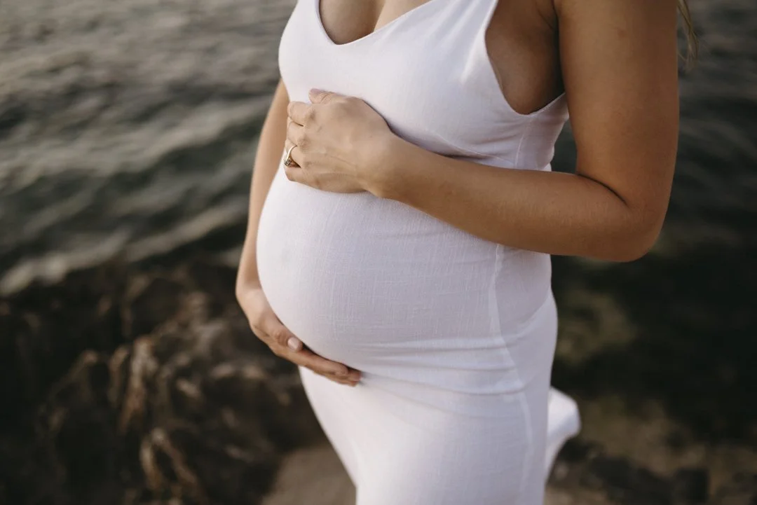 Pregnant woman in white dress holding her belly near water.