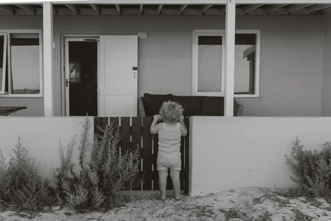 Black and white photo of a toddler standing at a low fence, looking towards a porch with an open door and windows.