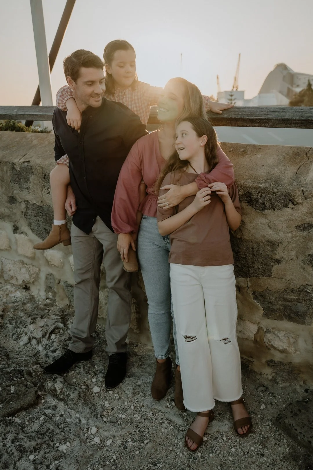 A family of four standing by a stone wall, enjoying a sunny day. The father is wearing a dark shirt, the mother a pink blouse, and the children are wearing casual outfits. Everyone is smiling and interacting warmly.