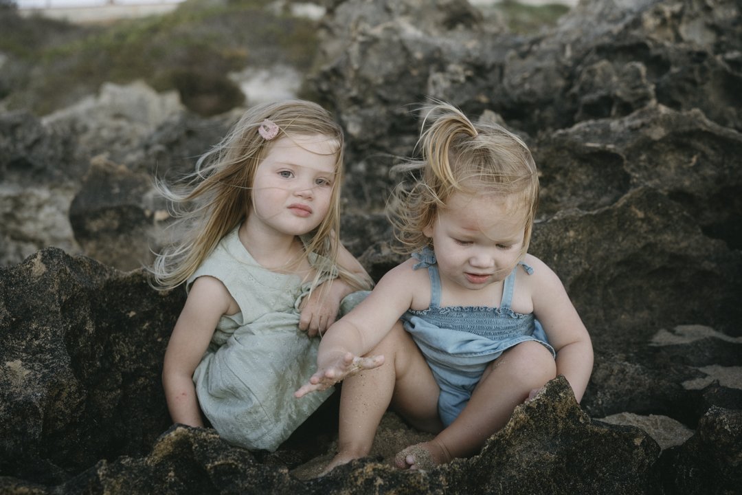 Two young children sitting on rough, rocky terrain, wearing light-colored clothes with their hair blowing in the wind.