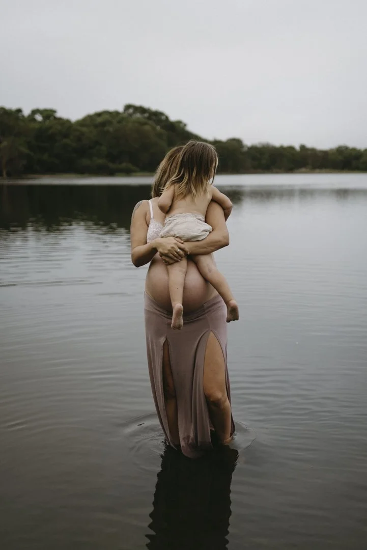 A woman standing in a lake holding a child, surrounded by trees.