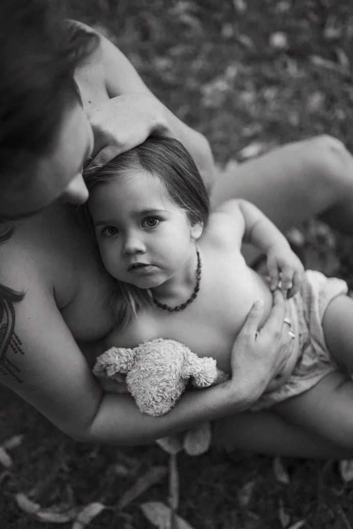 Black and white photo of a child wearing a necklace, being held by an adult. The child is holding a stuffed animal.