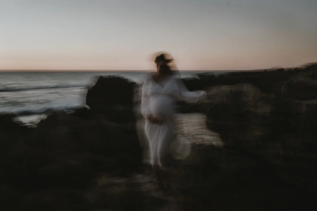 Blurred silhouette of a person in white standing near rocks by the sea at sunset.