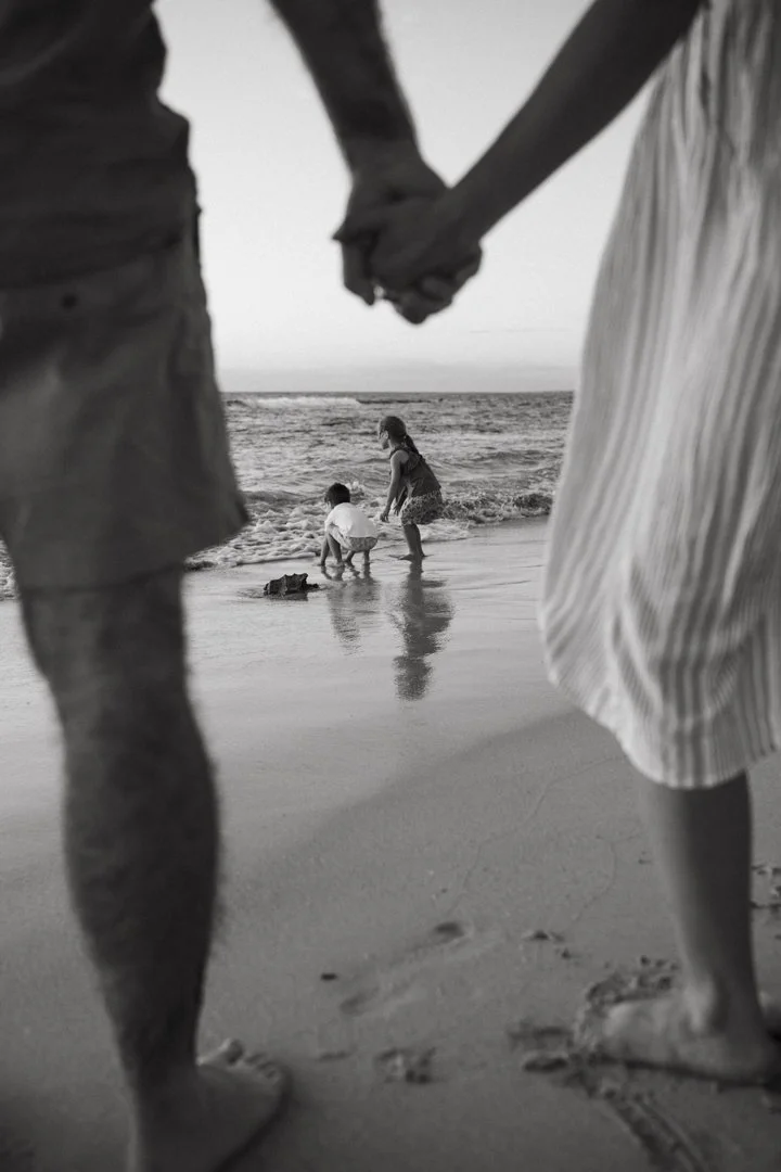 Black and white photo of a family at the beach, two adults holding hands in the foreground, two children playing by the shore.