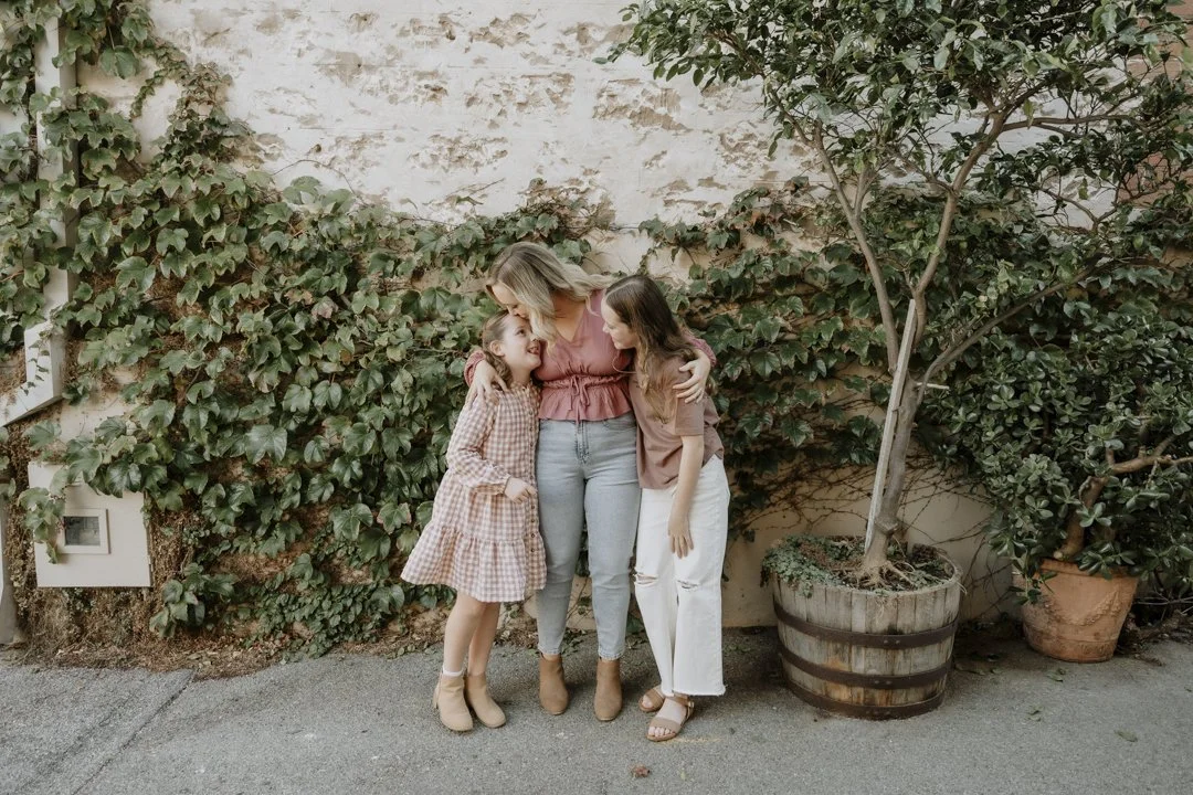 Three people embracing in front of an ivy-covered wall, with two potted trees nearby.