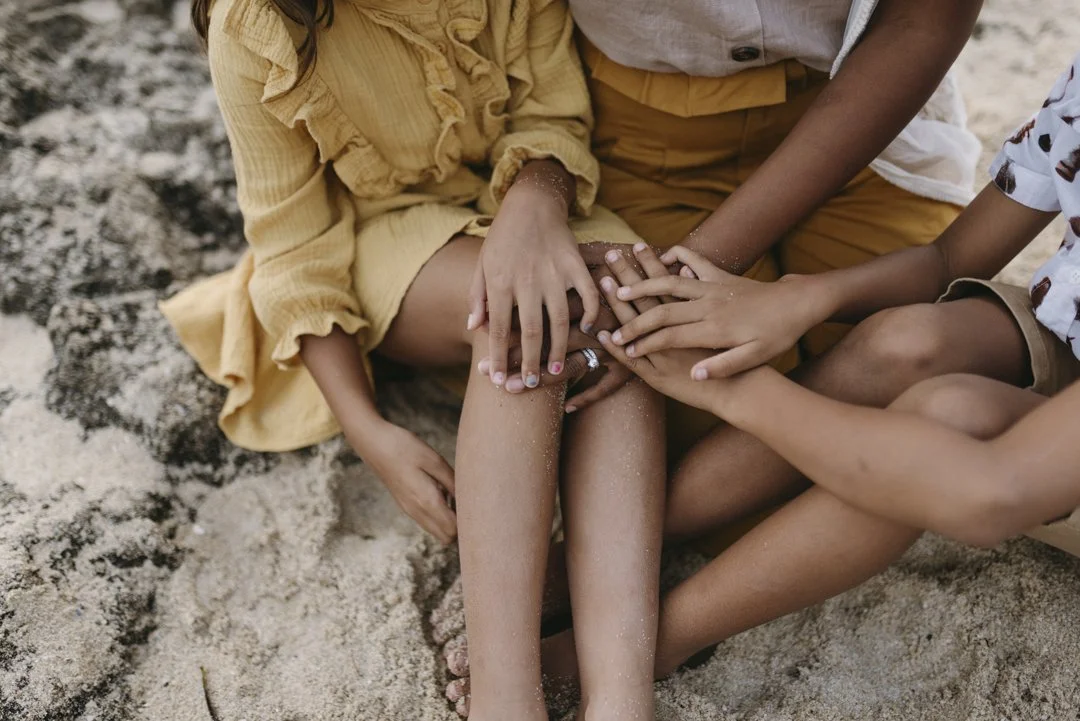 Close-up of children sitting on sandy beach, hands together in gesture of unity, wearing yellow clothing.