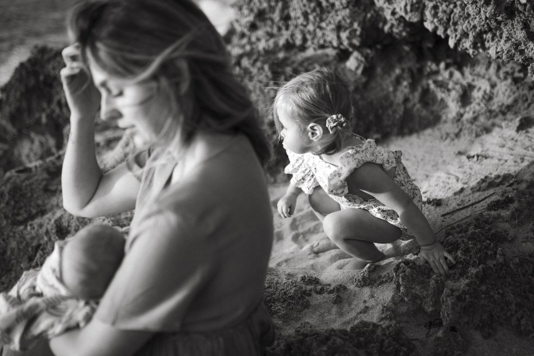 Woman holding a baby and a young child near rocks and sand, black and white photo.
