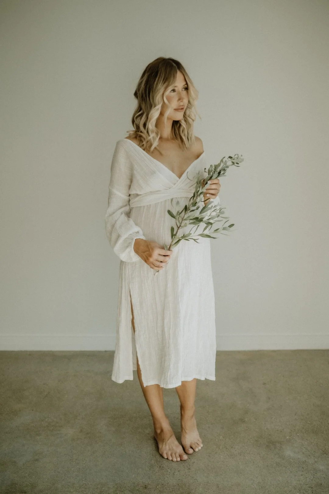 Woman in a white off-shoulder dress holding a branch with green leaves, standing barefoot on a concrete floor against a plain background.